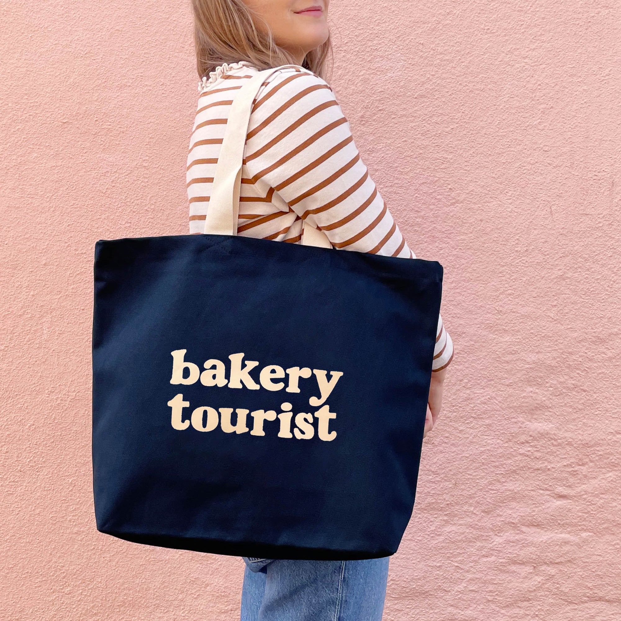Person holding a navy tote bag with 'bakery tourist' text against a pink background